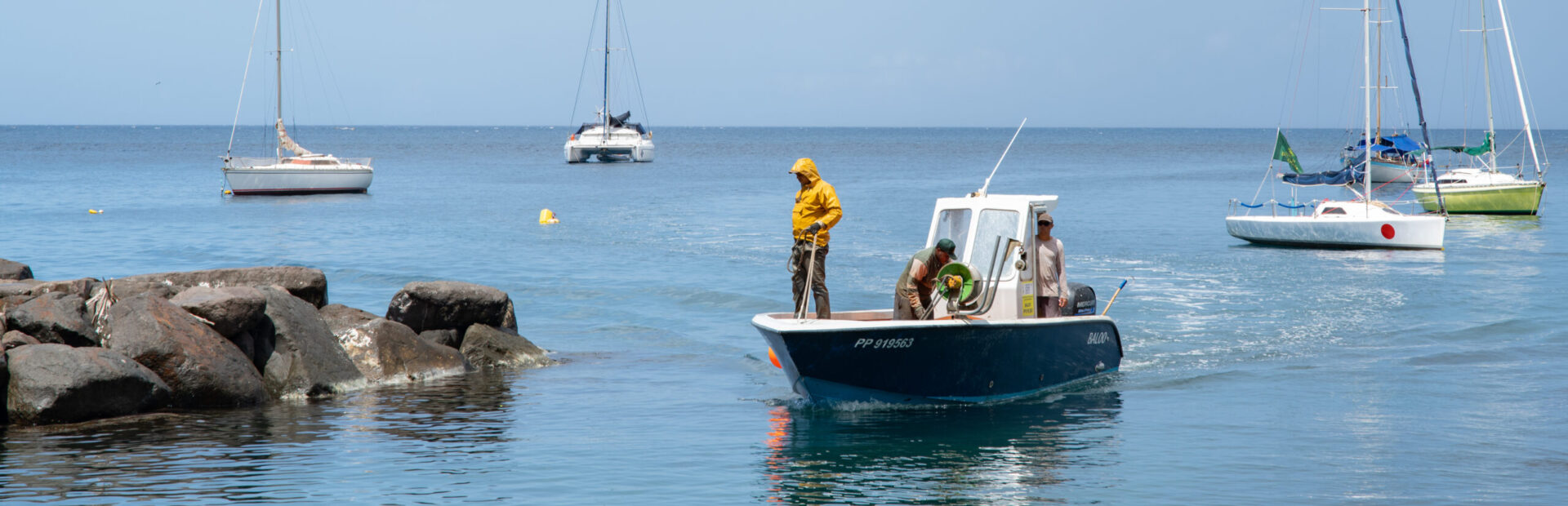 Trois pêcheurs antillais sur une petite embarcation bleue en mer, entourés de voiliers, au large des côtes.