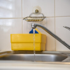 Robinet d’évier de cuisine avec filet d’eau coulant, sur fond de carrelage blanc et bac de rangement jaune.